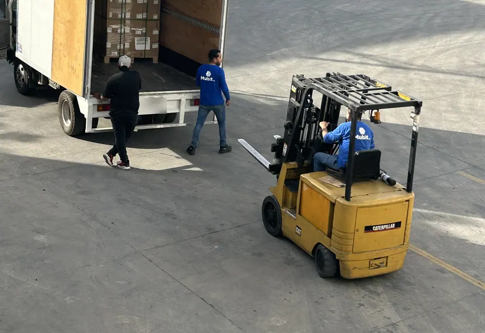 Montacargas amarillo y dos operadores de Mubit junto a un camión de caja con tarimas de cajas