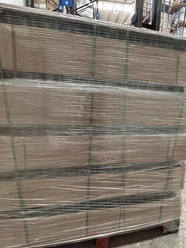 Tarima de cajas de cartón con fleje verde y película plástica, con racks de tarimas al fondo
