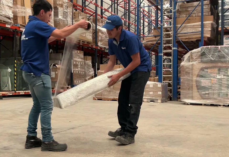 Dos operadores de Mubit con uniforme azul desenrollando película emplayable frente a los racks